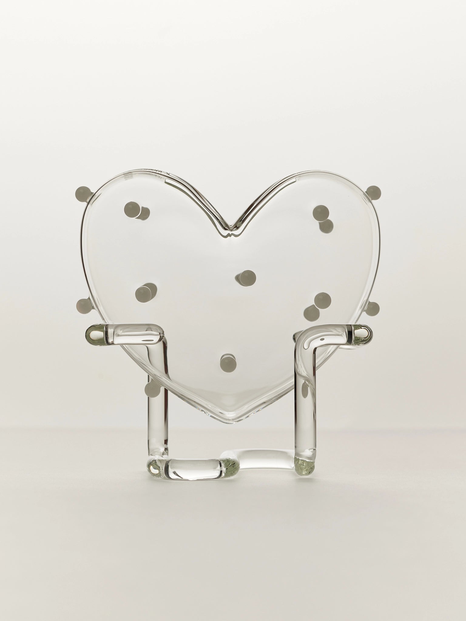 A clear glass heart-shaped vase with white glass balls on a clear stand against a light background